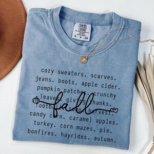 Op de afbeelding: Een lichtblauw Comfort Colors t-shirt met het woord "fall" in cursief en een lijst met herfstthemawoorden. Een gouden ketting met een hanger is over het shirt gedrapeerd.