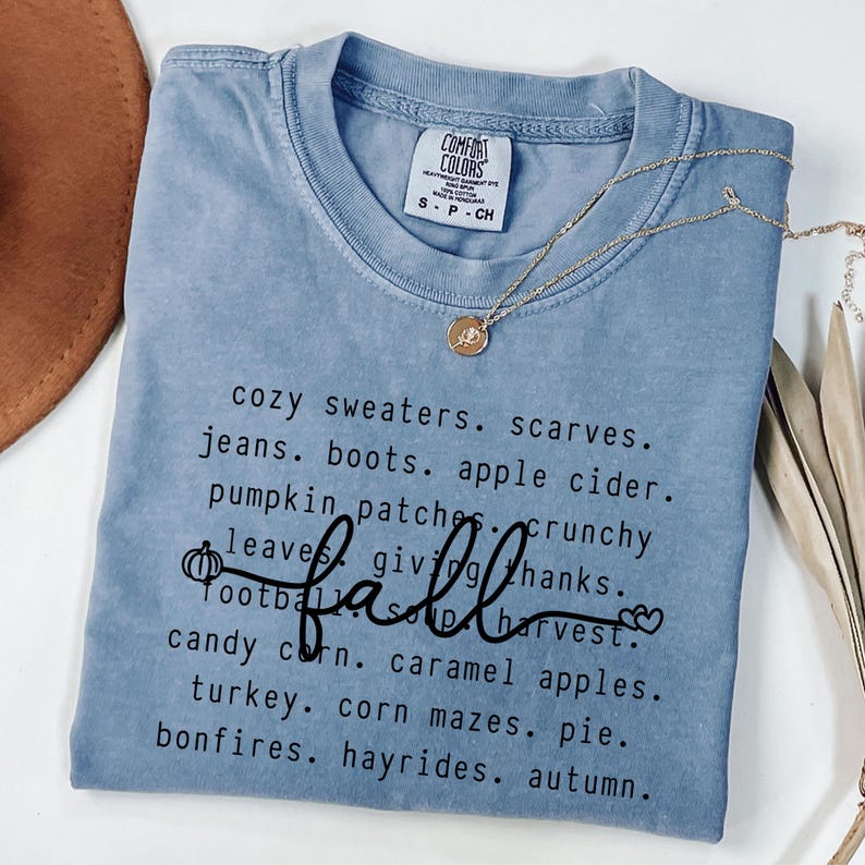 Op de afbeelding: Een lichtblauw Comfort Colors t-shirt met het woord "fall" in cursief en een lijst met herfstthemawoorden. Een gouden ketting met een hanger is over het shirt gedrapeerd.
