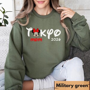 Pode incluir: Camisola verde militar com o texto "Tokyo Mom 2026" em branco e vermelho. O design inclui um gr&aacute;fico da Minnie Mouse com um la&ccedil;o vermelho. A camisola &eacute; de cor s&oacute;lida e tem mangas compridas.
