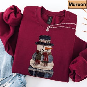 Op de afbeelding: Bordeaux sweatshirt met een sneeuwman applicatie. De sneeuwman heeft een zwarte hoed, een oranje wortelneus en een sjaal. Een zilveren ketting met een ronde hanger is over de trui gedrapeerd.
