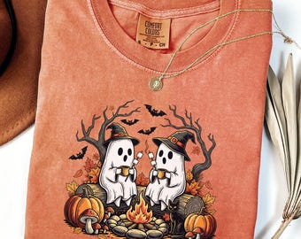 Può includere: T-shirt arancione Comfort Colors con grafica a tema Halloween. Il design include due fantasmi dei cartoni animati con cappelli da strega, seduti vicino a un falò, con zucche, pipistrelli e un gatto nero. Una collana d'oro è drappeggiata sulla maglietta.