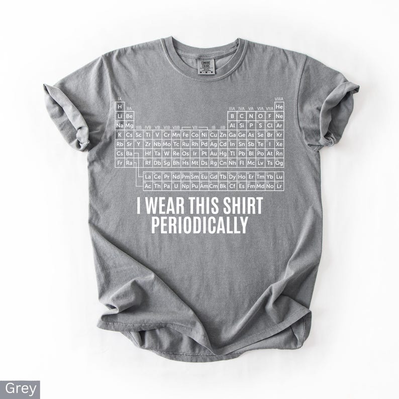 May include: A grey t-shirt with a white periodic table of elements design. The shirt has the text "I WEAR THIS SHIRT PERIODICALLY" printed below the table. The shirt is made of a soft material.