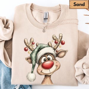 May include: A sand-colored crewneck sweatshirt featuring a graphic of a reindeer wearing a green Santa hat with a white pom-pom. The reindeer has a large red nose and antlers decorated with red and gold ornaments. The Gildan brand tag is visible.