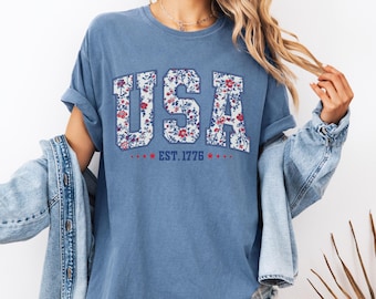May include: A blue t-shirt with the word "USA" in a floral pattern. The text "EST. 1776" is below the word "USA". The shirt has rolled-up sleeves and is worn with a denim jacket.