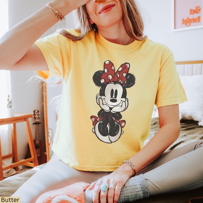 May include: A yellow t-shirt with a distressed Minnie Mouse graphic. The cartoon character has a red polka dot bow and is in a seated pose. The short-sleeved shirt is a casual style, suitable for everyday wear.