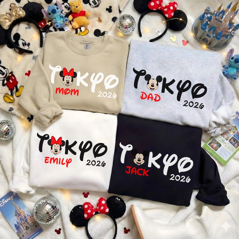 Pode incluir: Quatro camisolas de gola redonda bege, cinza, branca e preta, cada uma com um design "Tokyo 2026" com a cabe&ccedil;a do Mickey Mouse e os nomes "Mom", "Dad", "Emily" e "Jack". Acess&oacute;rios com tema da Disney rodeiam as camisolas.