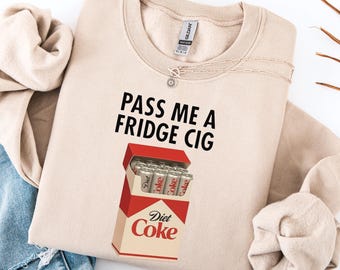 May include: A sand-colored sweatshirt with the text "PASS ME A FRIDGE CIG" in black. A graphic of a Diet Coke carton with Diet Coke cans is printed below the text. A silver necklace is visible.