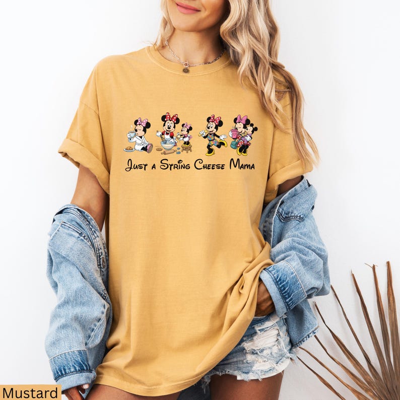 Op de afbeelding: Mosterdkleurig T-shirt met een cartoonafbeelding van Minnie Mouse-personages in verschillende poses. De tekst "Just a String Cheese Mama" staat onder de afbeelding. Het shirt heeft korte mouwen en een relaxte pasvorm.