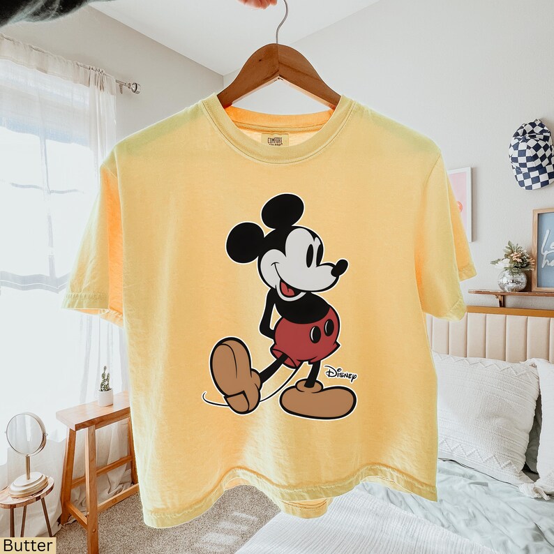 K&ouml;nnte beinhalten: Hellgelbes T-Shirt mit einer gro&szlig;en Mickey Mouse-Grafik. Die Zeichentrickfigur ist in Schwarz, Wei&szlig;, Rot und Braun gehalten. Das Disney-Logo ist in Schwarz unten rechts auf der Grafik aufgedruckt.