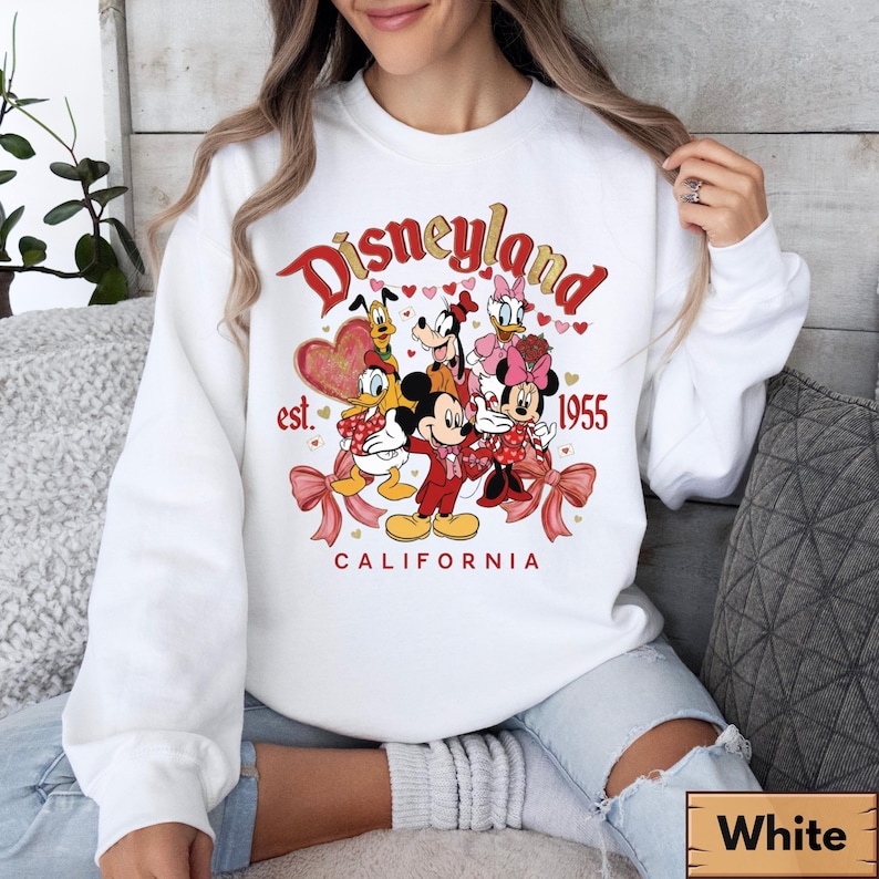 May include: White sweatshirt with a Disneyland graphic. The design features Mickey Mouse, Minnie Mouse, Donald Duck, Daisy Duck, Goofy, and Pluto. The text reads "Disneyland California est. 1955" in red and gold.