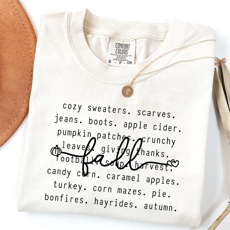 Op de afbeelding: Cr&egrave;mekleurig T-shirt met een lijst van herfstthemawoorden in het zwart, waaronder "fall" in cursief. Een gouden ketting met een hanger is over het shirt gedrapeerd. Het shirt heeft een label met "Comfort Colors".