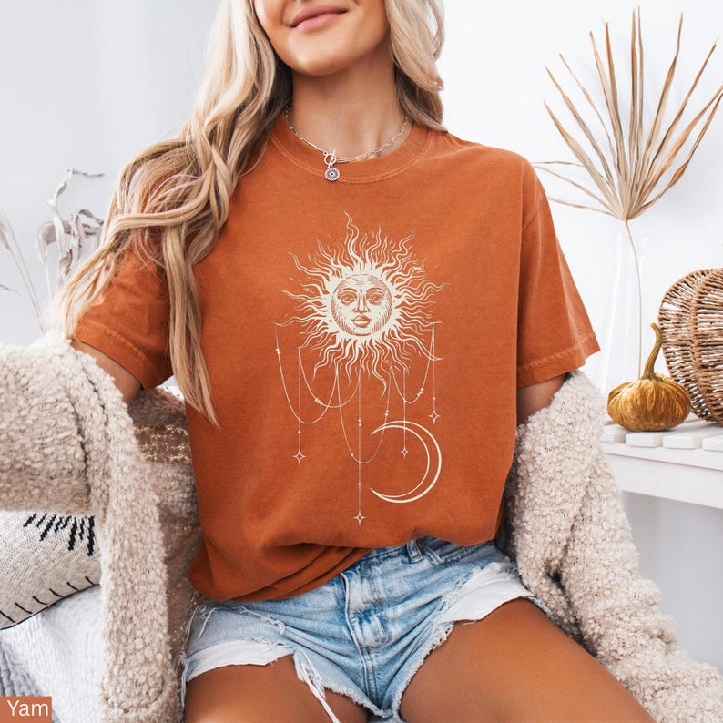 Puede incluir: Una camiseta de manga corta color naranja quemado con un estampado gr&aacute;fico blanco de un sol con cara, una luna creciente y cadenas de cuentas colgantes con detalles de estrellas. La camiseta tiene cuello redondo y shorts de mezclilla azul claro.