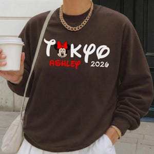 Pode incluir: Um moletom marrom com a palavra "Tokyo" em branco, um gr&aacute;fico da Minnie Mouse, o nome "Ashley" em vermelho e o ano "2026" em branco. O moletom tem mangas compridas.