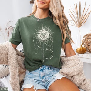Puede incluir: Una camiseta de manga corta color verde musgo con un estampado gr&aacute;fico en color crema que muestra un sol con rostro humano, una luna creciente y cadenas de cuentas colgantes con detalles de estrellas. Cuello redondo y corte holgado.