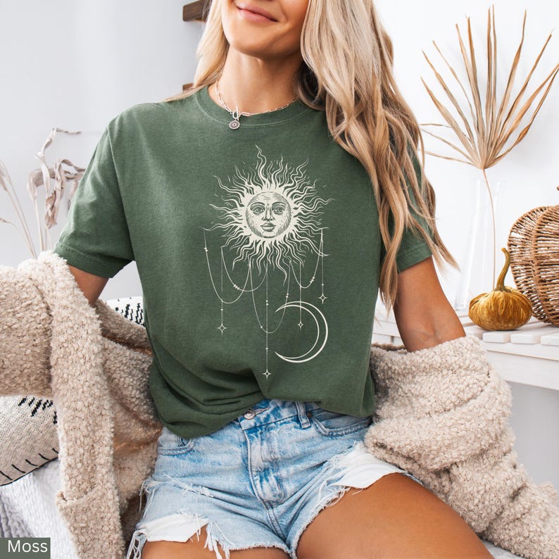 Puede incluir: Una camiseta de manga corta color verde musgo con un estampado gr&aacute;fico en color crema que muestra un sol con rostro humano, una luna creciente y cadenas de cuentas colgantes con detalles de estrellas. Cuello redondo y corte holgado.