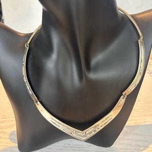 Könnte beinhalten: Eine goldfarbene Halskette mit einem V-förmigen Anhänger. Die Halskette wird auf einer schwarzen Schaufensterpuppe präsentiert. Der Anhänger hat ein geometrisches Design mit strukturierter Optik. Die Halskette ist im Choker-Stil.