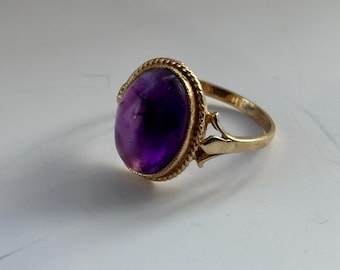 May include: A gold ring featuring a large, oval-cut amethyst gemstone. The deep purple stone is set in a gold band with a decorative, rope-like border. The ring's band has a delicate, ornate design on the sides.