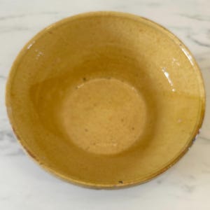 Pode incluir: Uma tigela de cerâmica redonda com um esmalte amarelo mostarda brilhante. A superfície interior apresenta uma textura subtilmente salpicada em todo o acabamento, com uma borda curva lisa e um design de bacia profunda e aberta, uniforme na cor.