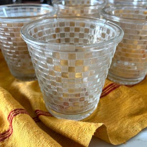 May include: A collection of clear glass tumblers with a woven basket weave pattern. The glasses are arranged on a yellow cloth with red stripes. The glasses are ideal for serving beverages.