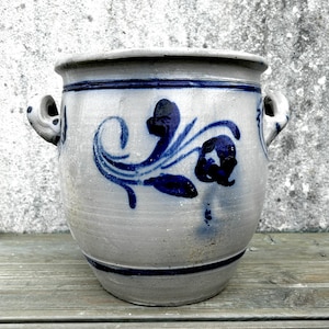 May include: A grey ceramic planter with blue floral patterns and two loop handles. The planter has a wide opening and a dark blue band around the top and bottom. The floral design is a stylized swirl with leaves and flower-like shapes.