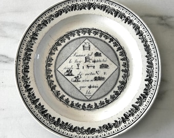 Peut inclure: Assiette en céramique blanche avec une bordure de vigne noire et un motif central en losange. Le losange contient du texte et des illustrations. L'assiette est ronde et a un bord légèrement surélevé.