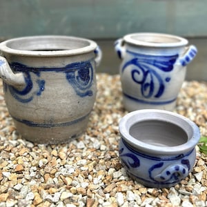 May include: Three ceramic pots with blue and white floral patterns. The pots are of varying sizes, with two featuring handles. They rest on a bed of small, light-coloured stones. The pots have a rustic, handmade appearance.