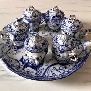 May include: A white ceramic serving tray with two handles featuring a blue floral pattern, holding five matching lidded cups. Each cup has a single handle and a lid with a small knob, decorated with the same blue and white floral motif.