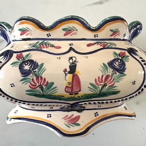May include: A cream-colored ceramic planter with a scalloped rim and handles. The exterior features hand-painted floral motifs in blue, green, and red, along with a central illustration of a person wearing a bonnet and apron, framed by blue lines.
