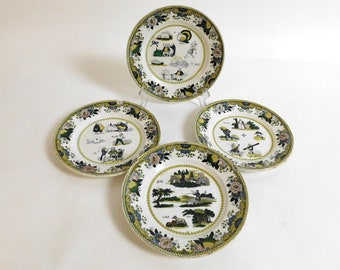 Peut inclure: Ensemble de quatre assiettes en céramique blanche avec des bordures florales vertes, jaunes et noires. Chaque assiette présente une scène différente de personnes dans un cadre rural.