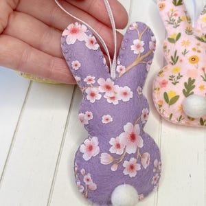 Pu&ograve; includere: Un ornamento di coniglio pasquale in feltro viola con una coda di pon pon bianca. Il coniglio &egrave; decorato con un motivo floreale rosa e bianco.