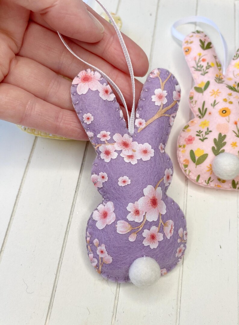 Pu&ograve; includere: Un ornamento di coniglio pasquale in feltro viola con una coda di pon pon bianca. Il coniglio &egrave; decorato con un motivo floreale rosa e bianco.