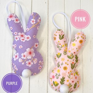 Pu&ograve; includere: Due ornamenti di coniglio in feltro con motivi floreali. Il coniglio a sinistra ha uno sfondo viola con fiori rosa e il coniglio a destra ha uno sfondo rosa con fiori gialli. Il testo "PINK" &egrave; in un cerchio rosa sopra il coniglio a destra e il testo "PURPLE" &egrave; in un cerchio viola sotto il coniglio a sinistra.