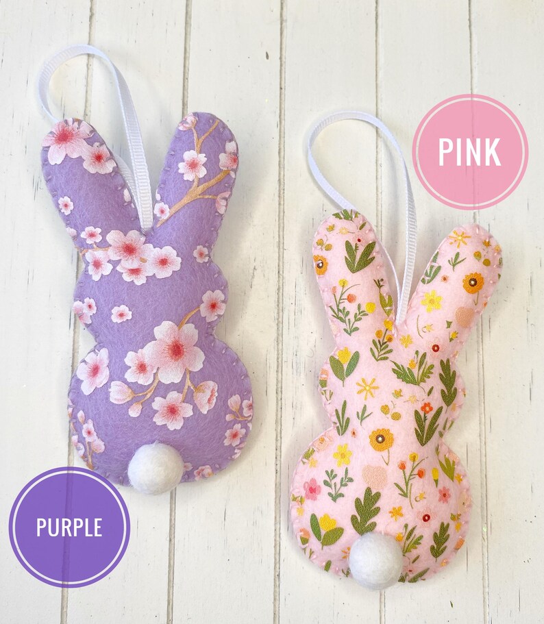 Pu&ograve; includere: Due ornamenti di coniglio in feltro con motivi floreali. Il coniglio a sinistra ha uno sfondo viola con fiori rosa e il coniglio a destra ha uno sfondo rosa con fiori gialli. Il testo "PINK" &egrave; in un cerchio rosa sopra il coniglio a destra e il testo "PURPLE" &egrave; in un cerchio viola sotto il coniglio a sinistra.
