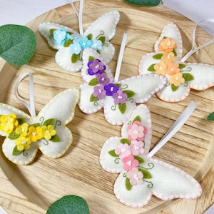 May include: Five felt butterfly ornaments with embroidered flowers in various colors, including yellow, pink, purple, and blue. The butterflies are hanging from white ribbon.