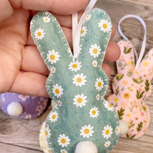Pu&ograve; includere: Un ornamento di coniglio in feltro verde con un motivo a margherita bianca. Il coniglio ha una coda a pompon bianco e un nastro bianco per appenderlo.