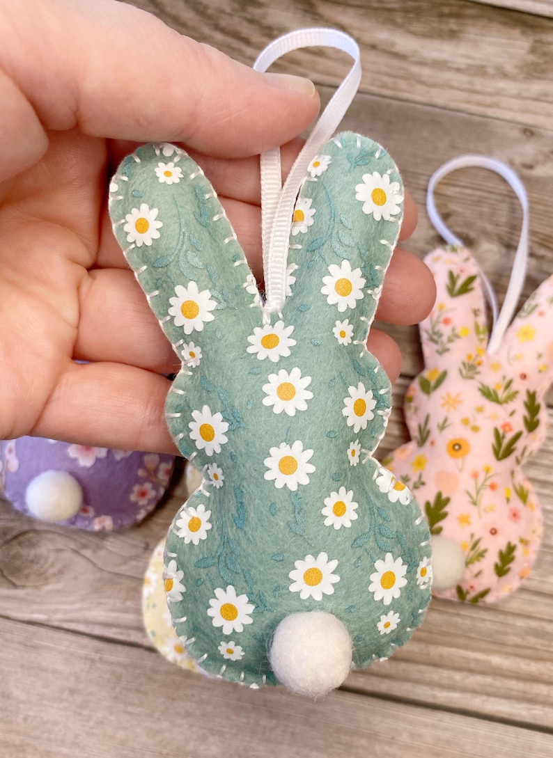 Pu&ograve; includere: Un ornamento di coniglio in feltro verde con un motivo a margherita bianca. Il coniglio ha una coda a pompon bianco e un nastro bianco per appenderlo.