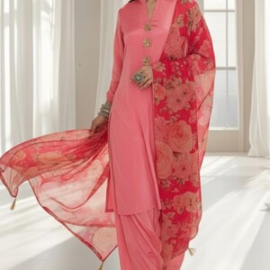 May include: A light pink salwar kameez ensemble. The outfit includes a long-sleeved tunic with decorative buttons, matching pants, and a sheer red floral-print scarf. The scarf has gold tassels. The outfit is worn by a person.