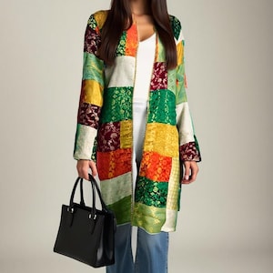 Op de afbeelding: Een patchworkjas in verschillende kleuren, waaronder groen, oranje, geel en rood. De jas wordt gedragen over een witte top en een blauwe spijkerbroek. Een zwarte handtas wordt in de hand gehouden.