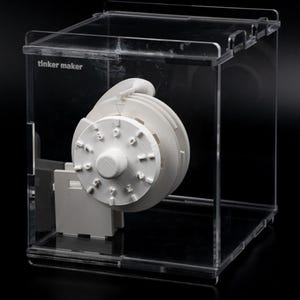 May include: A white, 3D-printed combination lock mechanism is encased in a clear acrylic box. The lock features a circular dial with numbers and a handle. The text "tinker maker" is printed on the box.