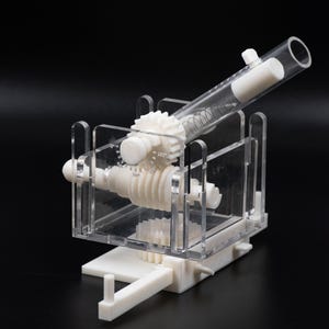 May include: A white and clear plastic mechanical device with gears, a spring, and a clear tube. The device is set against a black background. The gears are white and the clear tube is angled upwards.
