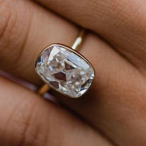Puede incluir: Primer plano de un anillo de oro con un gran diamante claro rectangular. El diamante está engastado en una banda de oro. El anillo está en el dedo de una persona.