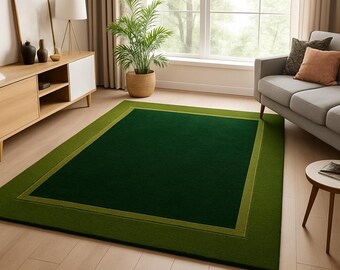 May include: A rectangular green area rug with a darker green center and a lighter green border. The rug is placed on a light-colored wooden floor in a living room setting. A sofa, side table, and cabinet are also visible.
