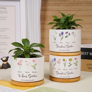 Könnte beinhalten: Drei weiße Keramik-Blumentöpfe mit Holzsockeln, die jeweils eine grüne Pflanze enthalten. Die Töpfe sind mit floralen Illustrationen und den Texten "Mom's Garden" und "Grandma's Garden" verziert. Der Satz "Love Grows Here" ist ebenfalls enthalten.