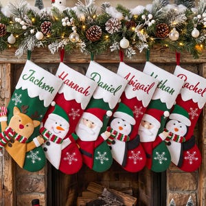 Op de afbeelding: Zes kerstsokken hangen op een rij, elk met een witte boord en een gepersonaliseerde naam: Jackson, Hannah, Daniel, Abigail, Nathan en Rachel. De sokken tonen feestelijke figuren zoals een rendier, een sneeuwpop en een pinguïn.