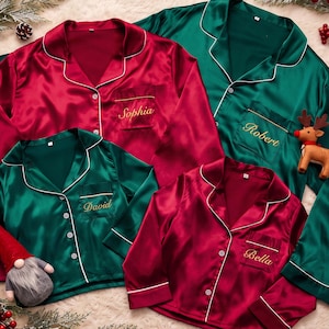 May include: A set of four long-sleeved button-down pajama shirts in red and green satin fabric with white piping. The shirts feature embroidered names: Sophia, Robert, David, and Bella. Each shirt has a chest pocket and a notched collar.