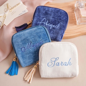 May include: Three square zippered pouches with textured fabric. The dark blue pouch features the name Abigail, the medium blue pouch features the name Maggie, and the white pouch features the name Sarah. Each pouch includes a matching tassel zipper pull.