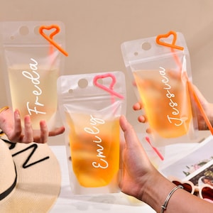 May include: Three clear plastic drink pouches filled with colorful beverages. Each pouch has a heart-shaped straw and a name written on it: Freda, Emily, and Jessica. A straw and a sun hat are also visible.