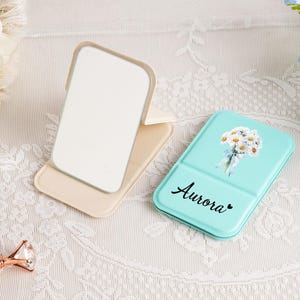 May include: A beige folding mirror and a light blue compact with a floral design and the name "Aurora" in script. The items are on a white lace tablecloth, with a pen and a diamond-shaped object in the foreground.
