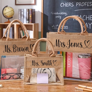Può includere: Tre borse tote in iuta con tasche frontali trasparenti. Ogni borsa ha un manico e un nome stampato in corsivo nero: "Ms. Brown", "Mrs. Smith" e "Mrs. Jones". Le borse sono piene di libri, un bicchiere e altri oggetti.