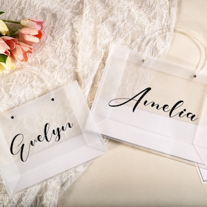 May include: Two white gift bags with clear panels, each bearing a name in elegant black script: "Evelyn" and "Amelia." The bags have white handles and are set against a backdrop of lace and a few pink and yellow tulips.